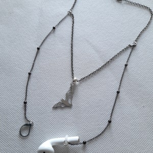 May include: A black chain necklace with a bat pendant and black beads. The necklace is attached to a pair of white earbuds with a black hook.
