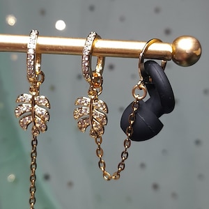 May include: Gold-toned earrings with leaf-shaped pendants, encrusted with small, clear stones. One earring has a chain that dangles below the leaf. A pair of black earbud covers are attached to a gold chain. The earrings hang from a gold bar.