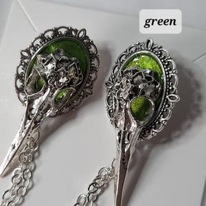 May include: A pair of silver tone earrings with a bird skull design. The skull is set in a filigree frame with a green enamel fill. The earrings are attached to a silver chain.