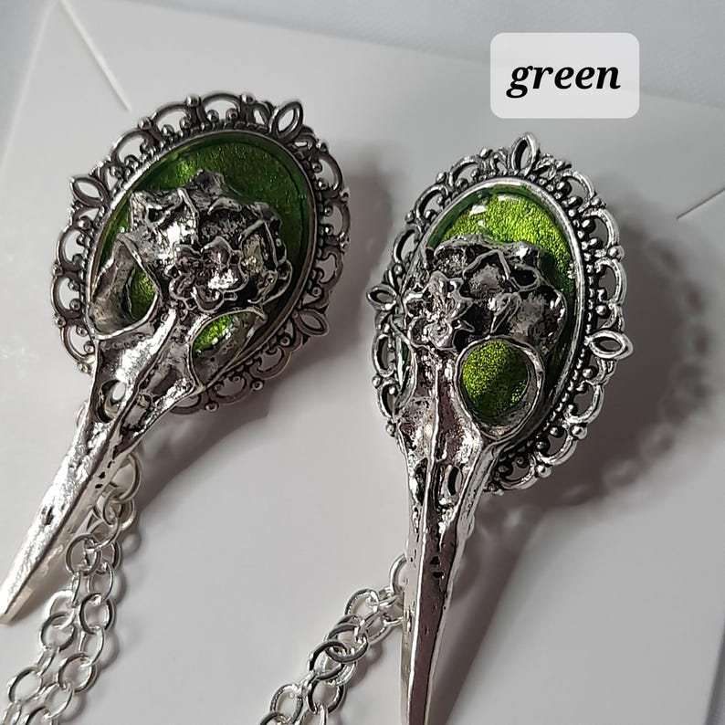 May include: A pair of silver tone earrings with a bird skull design. The skull is set in a filigree frame with a green enamel fill. The earrings are attached to a silver chain.