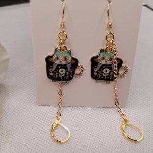 May include: A pair of gold-colored dangle earrings. Each earring features a black teacup with a cat design. The cat has blue and yellow stripes. The teacup has a gold handle and is decorated with flowers. The earrings have a gold chain and a clasp.