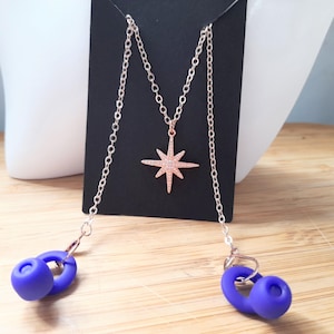 May include: A rose gold necklace with a starburst pendant and two blue silicone earbud holders. The necklace hangs on a black display card. The earbuds are connected to the necklace by a chain.