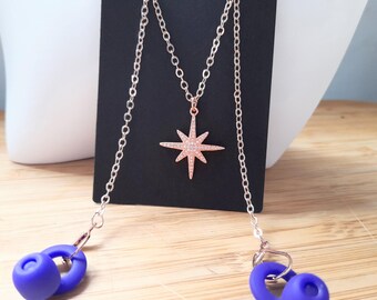 May include: A rose gold necklace with a starburst pendant and two blue silicone earbud holders. The necklace hangs on a black display card. The earbuds are connected to the necklace by a chain.