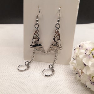 May include: Silver-toned bat earrings. Each earring features a bat silhouette pendant with a chain and a lever-back closure. The earrings are displayed on a white card, with a white flower in the background.