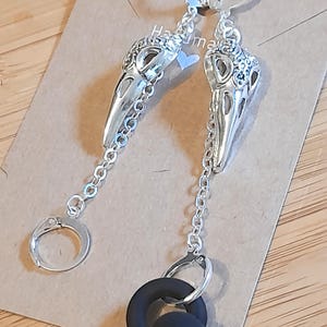 May include: A pair of silver-toned earrings featuring bird skull pendants suspended from chains. One earring includes a small silver hoop, while the other features a silver hoop attached to a black circular silicone ring. The earrings are displayed on a card.