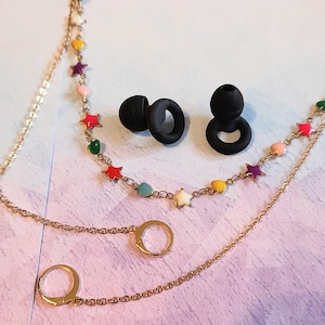 May include: A gold-colored necklace with a chain and a charm featuring a small ring. A second necklace has a chain with colorful star and heart charms. Two black earbud tips are also in the image.