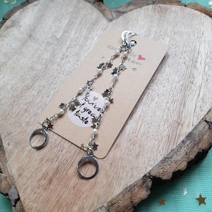 May include: Silver-toned earrings with a celestial theme. Each earring features a crescent moon at the top, a chain adorned with star charms and faux pearls, and a circular clasp. A card with the text "You've got great taste" is in the background.