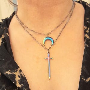 May include: A layered necklace with a dark chain. The top layer features a crescent moon pendant, and the bottom layer has a sword pendant. Both pendants have a rainbow iridescent finish, reflecting various colors.