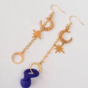 May include: Gold-colored celestial earrings featuring crescent moons and star charms. The earrings hang from gold-colored hooks and chains. A pair of dark purple earplugs are attached to the earrings.