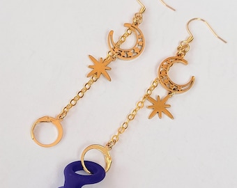May include: Gold-colored celestial earrings featuring crescent moons and star charms. The earrings hang from gold-colored hooks and chains. A pair of dark purple earplugs are attached to the earrings.