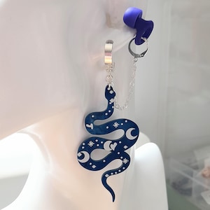 May include: A dark blue snake-shaped earring with white celestial designs, including moons and stars, hangs from a silver hoop. A silver chain connects the earring to a silver ring, and a purple ear plug is visible.