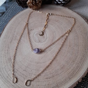May include: A gold chain necklace with a purple stone pendant and a crescent moon charm. The necklace is layered and has two chains.