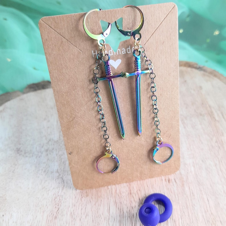 May include: A pair of iridescent sword earrings with matching handcuff charms. The earrings are attached to lever-back closures and hang from a brown card with the text "Handmade" and a white heart. A small blue object is at the bottom.