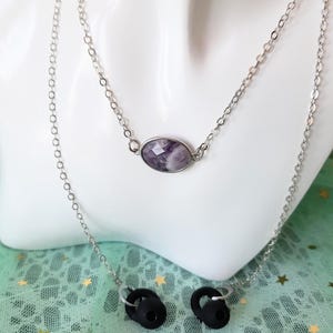 May include: A silver-toned metal chain necklace designed to hold wireless earbuds. The chain features a central oval-shaped faceted purple and white stone pendant. Two black silicone earbud holders are attached to the ends of the silver chain.