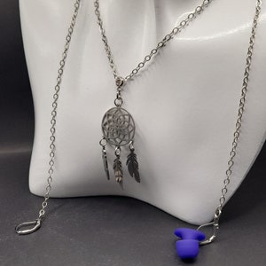 May include: Silver chain necklace with a dreamcatcher pendant and three feathers. The necklace is on a white mannequin bust. There are also two blue earplugs on the surface.
