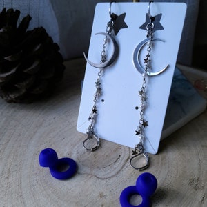 May include: A pair of silver crescent moon earrings with a dangling chain of silver stars and white beads. The earrings are displayed on a white card with a star design.