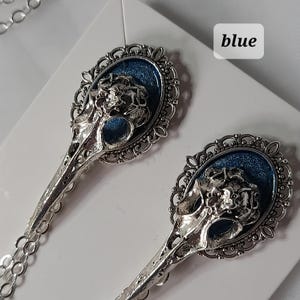 May include: A pair of silver earrings with blue enamel and a skull design. The earrings are in the shape of a bird's head with a detailed filigree design.