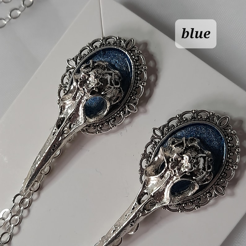 May include: A pair of silver earrings with blue enamel and a skull design. The earrings are in the shape of a bird's head with a detailed filigree design.