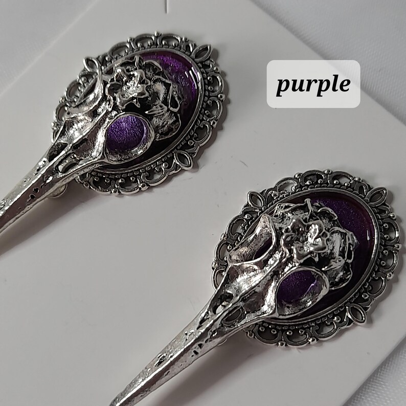 May include: A pair of silver earrings with a purple skull design. The skull is set in a filigree oval frame.