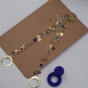 May include: Gold chain earrings with small leaf charms and colorful gemstones. The earrings are connected by a chain with a small circle at each end. The earrings are on a brown background with the text "Handmade" visible.