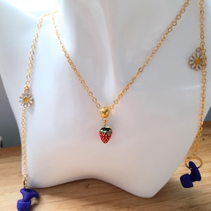 May include: A gold-colored necklace with a red strawberry pendant and daisy and blue charm accents. The necklace is displayed on a white mannequin bust. The necklace has a delicate chain design.