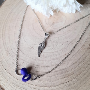 May include: A two-strand necklace set. The top strand features a silver-toned chain with a pendant shaped like a wing. The bottom strand has a silver-toned chain with a purple silicone object. The background is a wooden surface with a white flower.