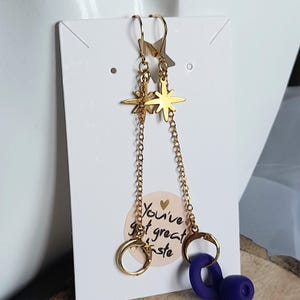 May include: Gold-coloured dangle earrings with star-shaped studs and chain details. Each earring has a gold hoop and a dark purple accent. The earrings are displayed on a white card with the text "You've got great taste."