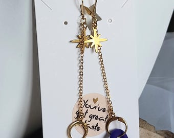 May include: Gold-colored dangle earrings featuring star-shaped studs and chain details. Each earring has a gold hoop and a dark purple accent. The earrings are displayed on a white card with the text "You've got great taste."