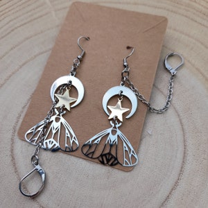May include: A pair of silver earrings with a crescent moon and star charm, and a moth charm. The earrings are connected by a silver chain.