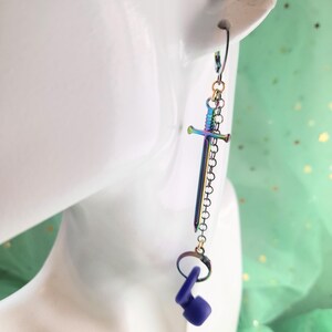 May include: A single earring featuring a sword design with a rainbow finish. The sword hangs from a chain and is connected to a purple object. The earring is displayed on a white mannequin head.