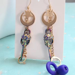 May include: A pair of gold-colored earrings with a celestial theme. Each earring features a crescent moon and star charm, connected to a bird skull pendant with iridescent coloring. The earrings dangle from gold-colored hooks.