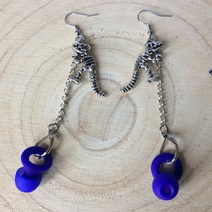 May include: A pair of dangle earrings featuring a silver dinosaur skeleton charm and a purple dangling charm.