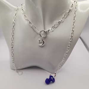 May include: A silver chain necklace with a crescent moon and star charm. The necklace is layered with a longer silver chain with a blue silicone earplug at the end.