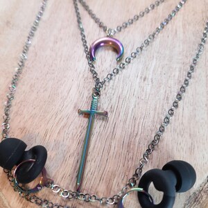 May include: A multi-layered necklace with a sword pendant, a crescent moon pendant, and a chain for earbuds. The pendants and chains have a metallic, iridescent finish. The earbuds are black.