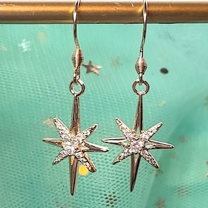 May include: A pair of gold-colored starburst earrings. Each earring features a starburst design with multiple points, and is adorned with small, sparkling crystals. The earrings hang from a simple hook.