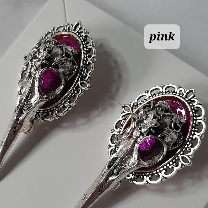 May include: A pair of silver earrings with a pink skull design. The skulls are surrounded by a decorative filigree frame. The earrings are labeled "pink".