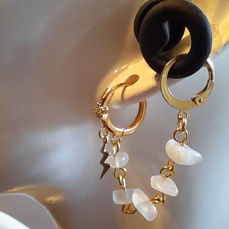 May include: A gold-toned hoop earring featuring a small star charm and a lightning bolt charm, connected by a delicate gold chain adorned with several small, smooth, white, translucent, irregular-shaped stone beads that drape elegantly.