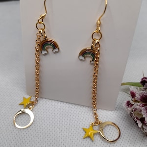 May include: A pair of gold earrings with a rainbow charm and a star charm hanging from a chain. The rainbow charm is multi-colored and has a white outline. The star charm is yellow.