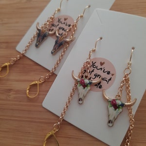 May include: Gold-tone earrings featuring a bull skull pendant with floral accents. The earrings have a chain design and lever-back closures. The text on the card reads "You've got great taste."