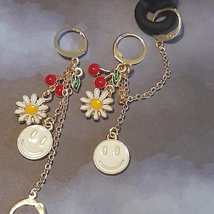 May include: Gold-tone earrings with a chain design. Each earring features a white daisy with a yellow center, red cherry accents, and a white smiley face charm. The earrings have a lever back closure.