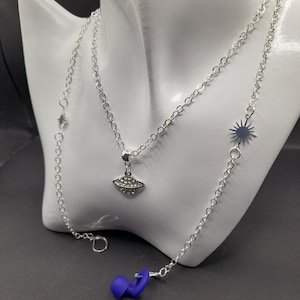 May include: A silver chain necklace with a small, silver charm of a planet with rhinestones. The necklace is layered on top of a longer silver chain with a star charm. A pair of blue earplugs are in the foreground.