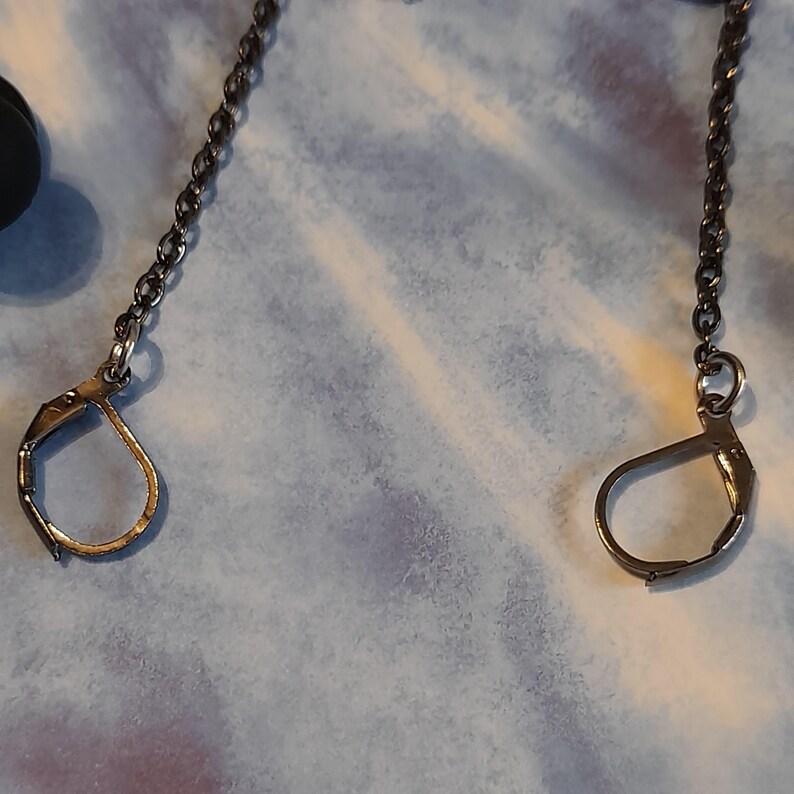 May include: Close-up of two silver-toned leverback earring clasps with attached chain. The clasps are open, revealing their design. The chains are made of small, dark metal links. The background is a light blue and white fabric.