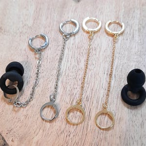 May include: A selection of hoop earrings, two in gold and one in silver, each with a chain and handcuff charm. Black earplugs are positioned on either side. The earrings are displayed on a wooden surface.