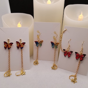 May include: Butterfly earrings in various colors, including red, orange, blue, and yellow. The earrings have a gold chain and a flower-shaped charm. The butterfly wings are detailed with intricate patterns. The earrings are displayed on white cards.