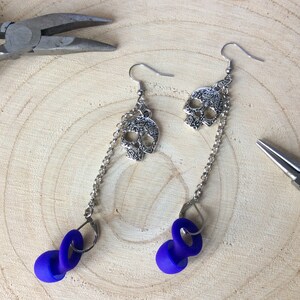 May include: A pair of dangle earrings with silver skull charms and purple resin beads. The earrings are on a wooden surface.