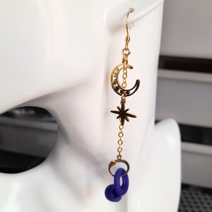 This image shows the loop earplug earings hanging from a model prop of the side of a face and ear. The earring charms hang down from the earhook and the chain with the clip at the end holds a loop earplug. This image shows an example of how the earring will look when the wearer is using the chain to hold the earplugs in readiness to use them in their ears.