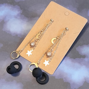 Puede incluir: Pendientes colgantes dorados con motivos celestiales, con amuletos de sol, luna y estrella. Los pendientes cuelgan de delicadas cadenas y se exhiben en una tarjeta marr&oacute;n. Tambi&eacute;n hay puntas de auriculares negras en la imagen.