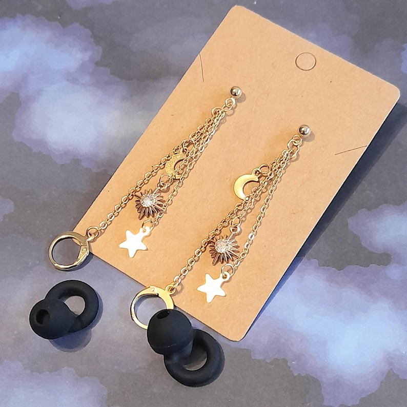 Puede incluir: Pendientes colgantes dorados con motivos celestiales, con amuletos de sol, luna y estrella. Los pendientes cuelgan de delicadas cadenas y se exhiben en una tarjeta marr&oacute;n. Tambi&eacute;n hay puntas de auriculares negras en la imagen.