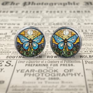 May include: A pair of round glass cabochons with silver settings featuring a stained glass butterfly design. The butterflies are blue, yellow, and green with a white moon in the background.