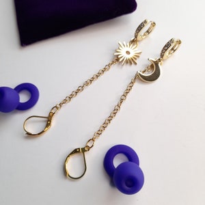 May include: A pair of gold earrings with a sun and moon design. The earrings are connected by a gold chain. The earrings are on a white surface.